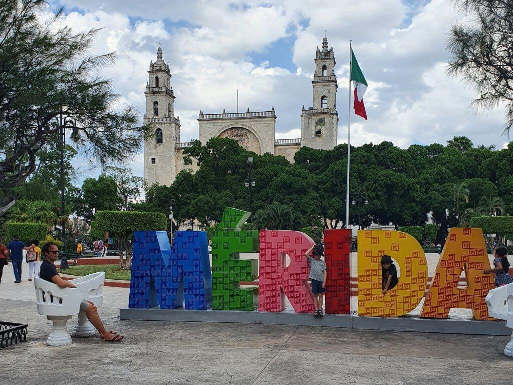 Kinderen poseren bij de kleurrijke Letras Turísticas van Mérida met de kerk op de achtergrond tijdens een vakantie in Mexico met kinderen